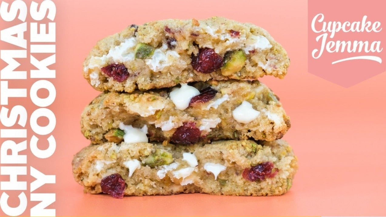 Three stacked Christmas cookies with white chocolate chips, cranberries, and pistachios on a pink background with recipe ingredients text