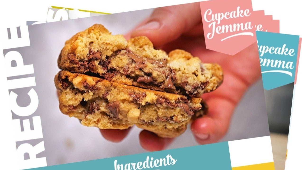 Close-up of a chunky chocolate chip cookie held over layered colorful recipe cards for New York Cookies bundle by Cupcake Jemma