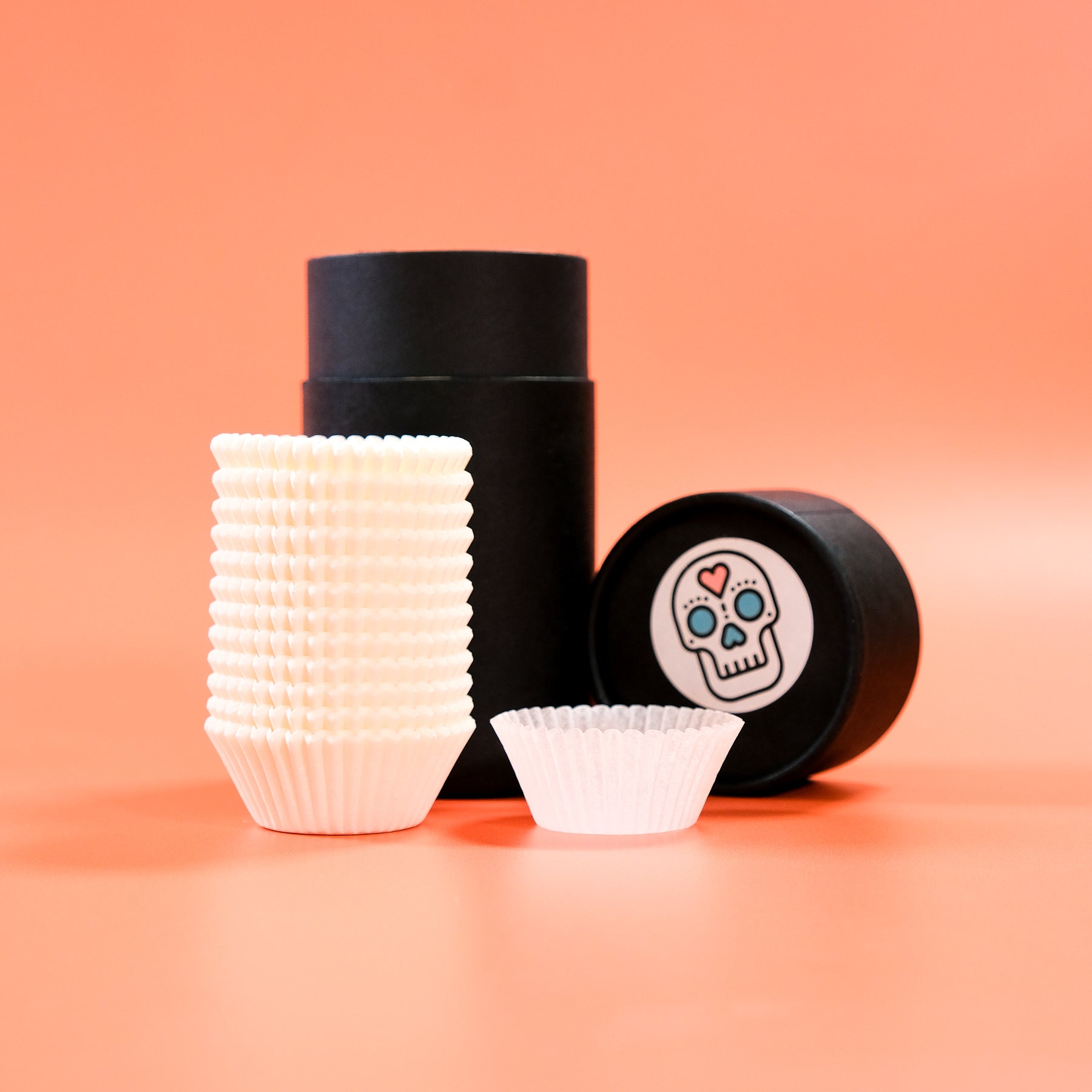 Stack of white mini cupcake cases with black cylindrical container on peach background for baking