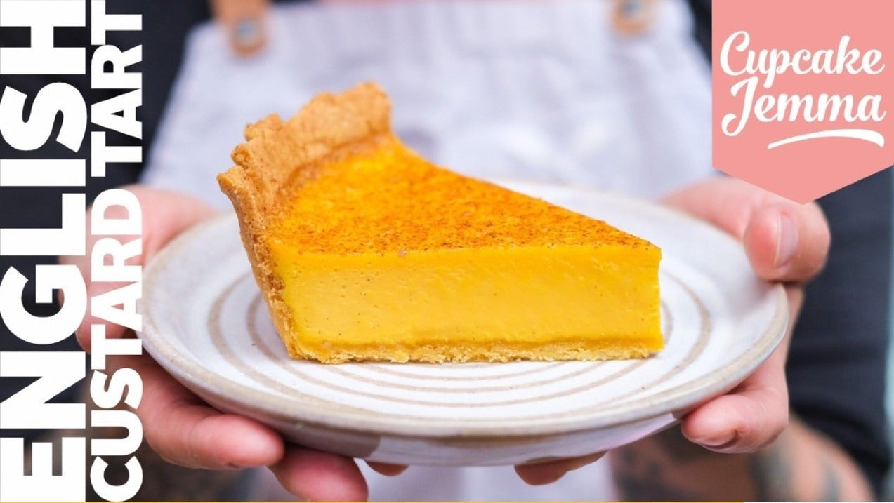 Slice of English custard tart on a plate held by hands, with Cupcake Jemma recipe details in background