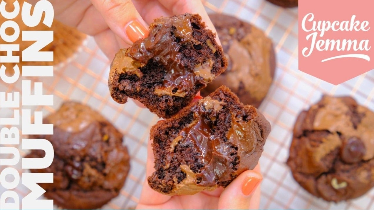 Hand holding a freshly baked double chocolate muffin broken in half showing gooey chocolate inside with more muffins in the background