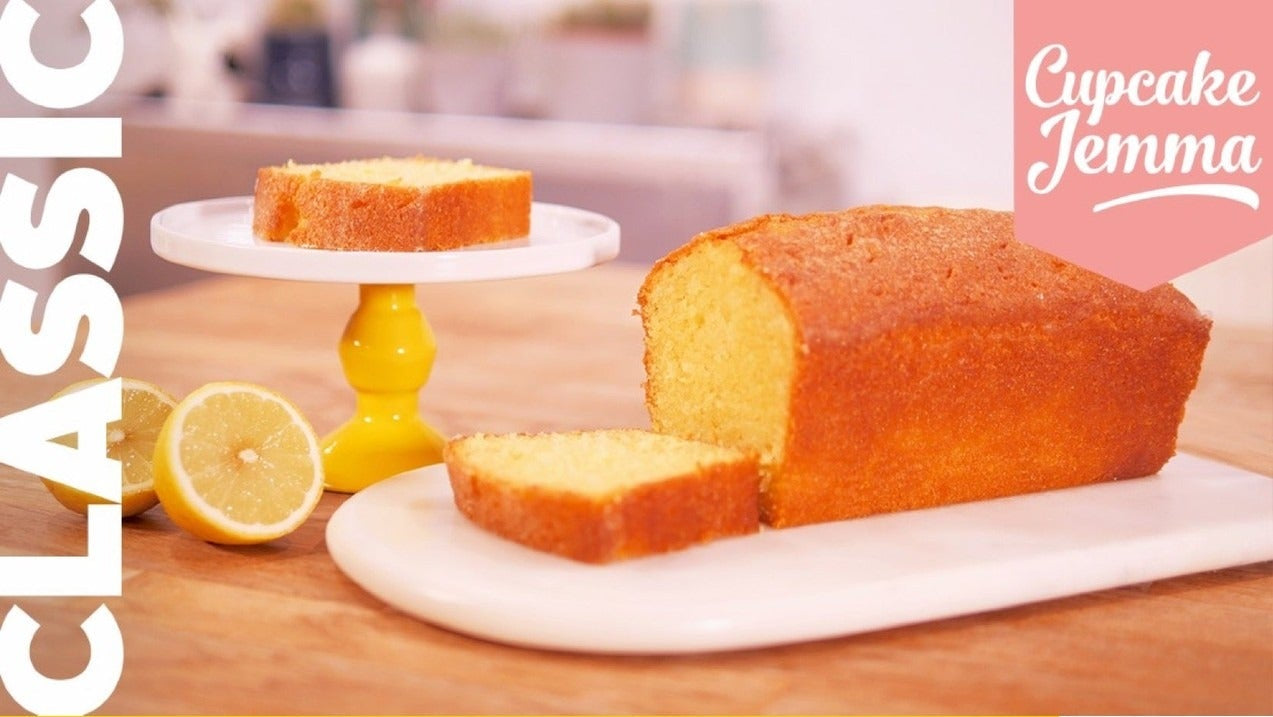 Lemon Drizzle Loaf Cake with a slice cut showing moist texture alongside fresh lemons on a wooden surface by Cupcake Jemma recipe