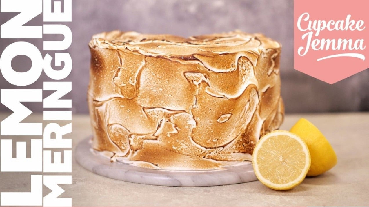 Lemon Meringue Cake with toasted meringue frosting and fresh lemon halves beside recipe ingredients by Cupcake Jemma