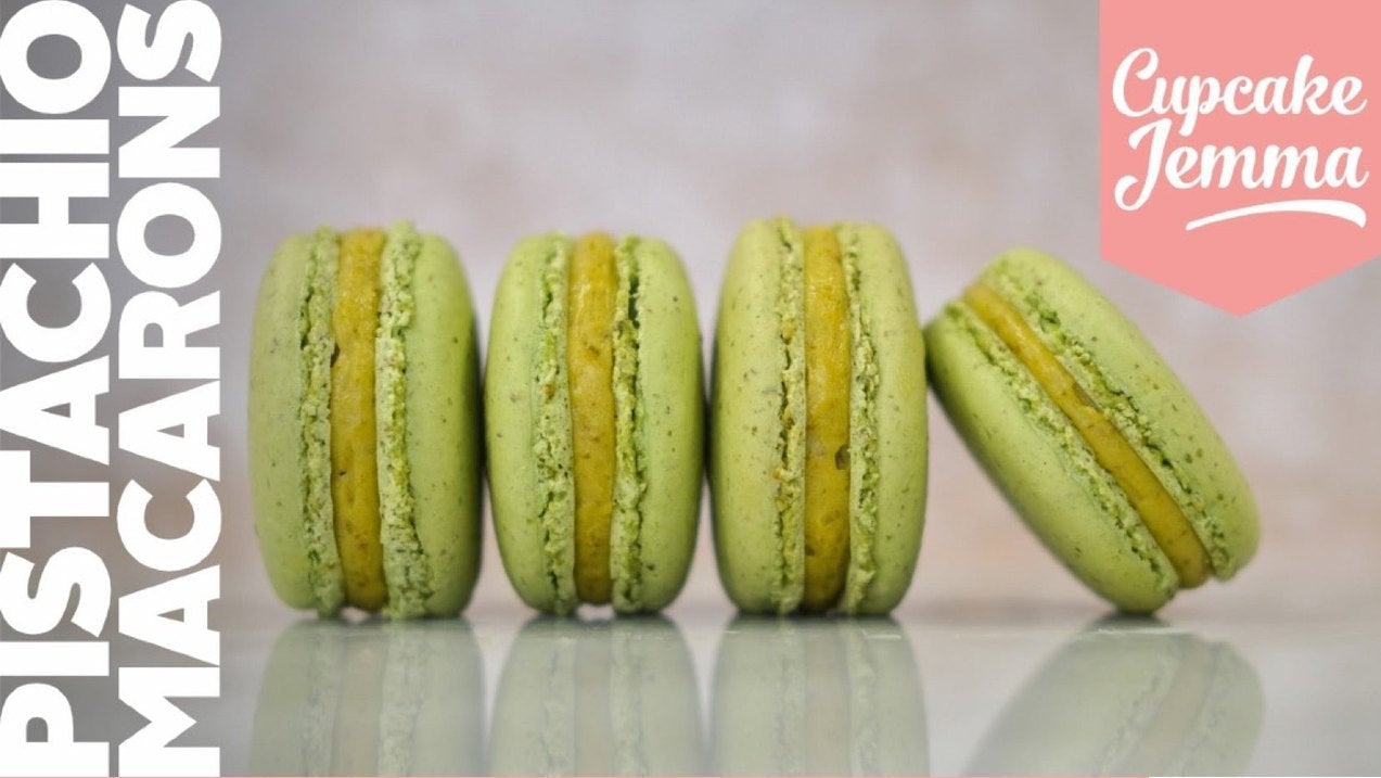 Four green pistachio macarons arranged in a row with recipe ingredients below on a pink background