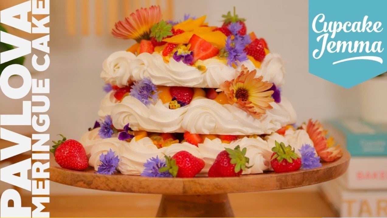 Three-layer Summer Meringue Pavlova Cake with cream, fresh fruit, and edible flowers on wooden stand by Cupcake Jemma
