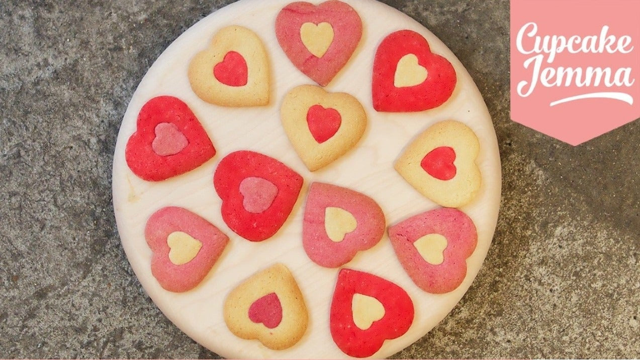Heart-shaped Valentine's cookies in red and pink on a wooden board with baking ingredients listed below for a downloadable recipe