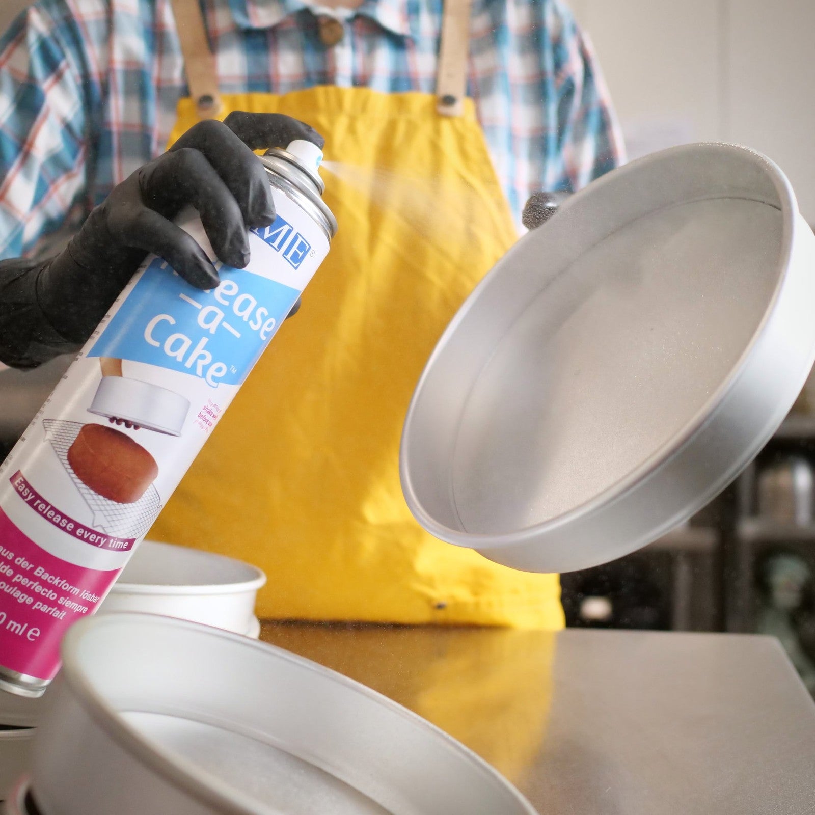 Person in yellow apron spraying Release A Cake Spray evenly on a round cake tin for baking preparation