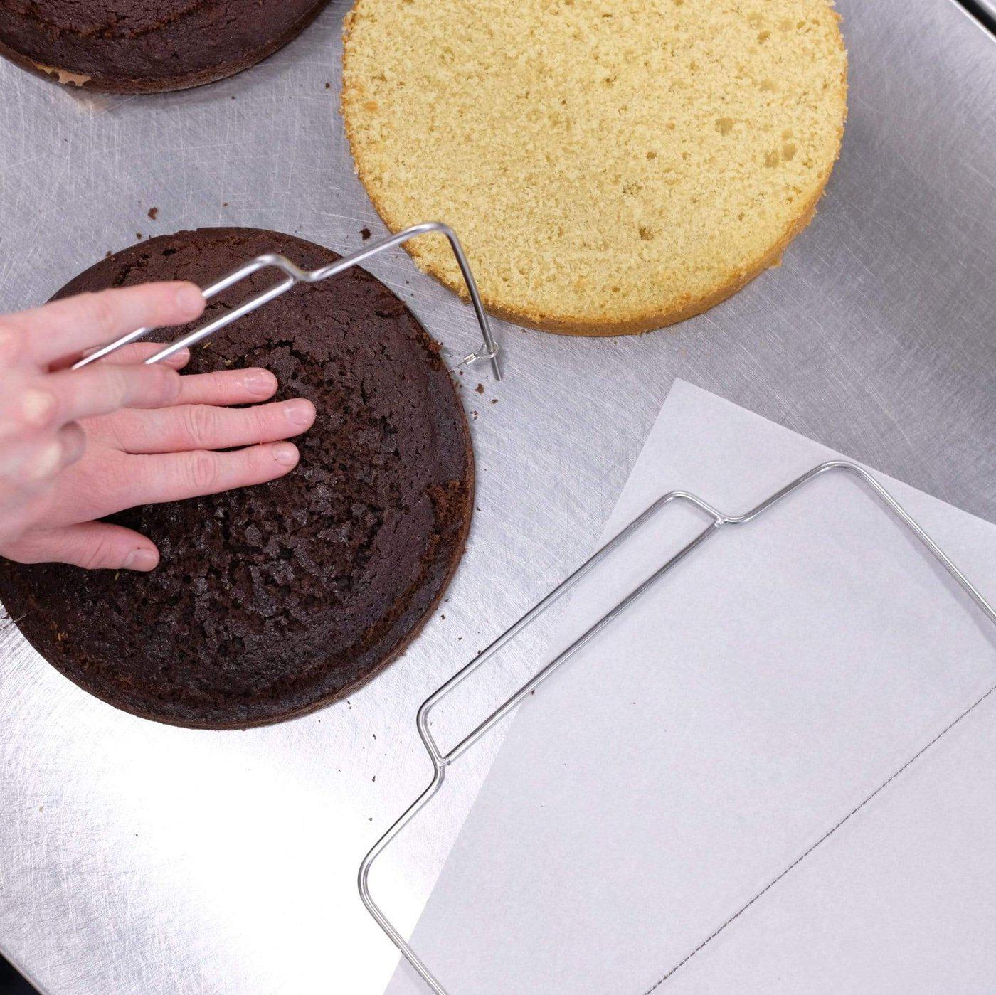 Cake Leveller being used to evenly slice a chocolate cake layer beside a vanilla cake on a baking surface