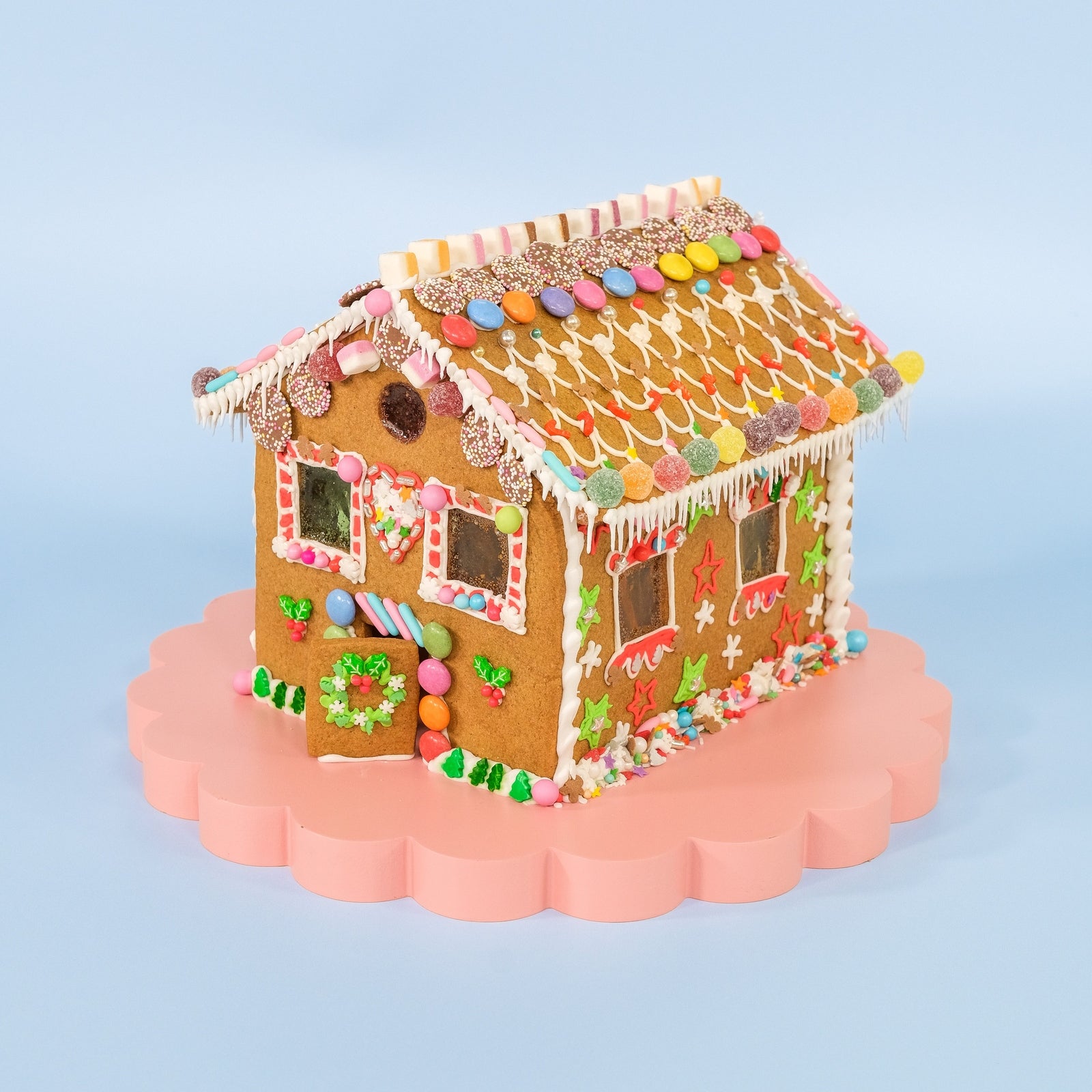 Decorated gingerbread house made using Gingerbread House Baking & Decorating Kit on pink stand against light blue background