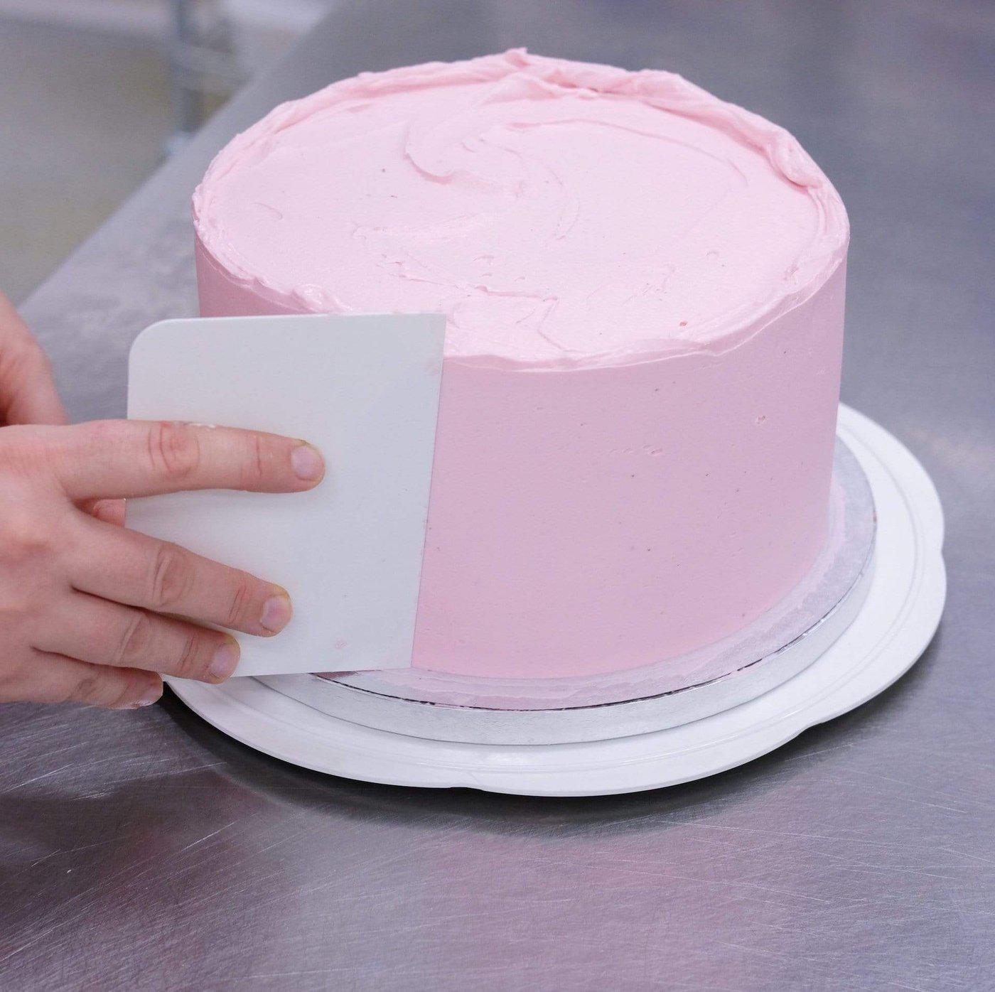 Cake Decorating Turntable with smooth pink frosted cake being evenly scraped for clean edges on a stainless steel surface