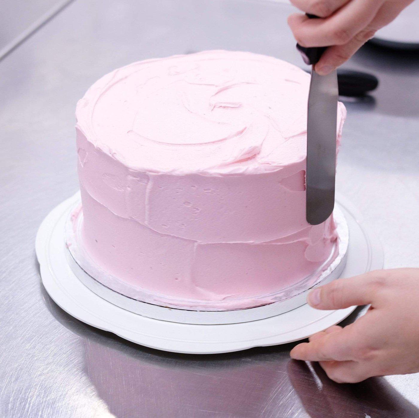 Hands smoothing pink frosting on a cake using a Cake Decorating Turntable on a stainless steel countertop