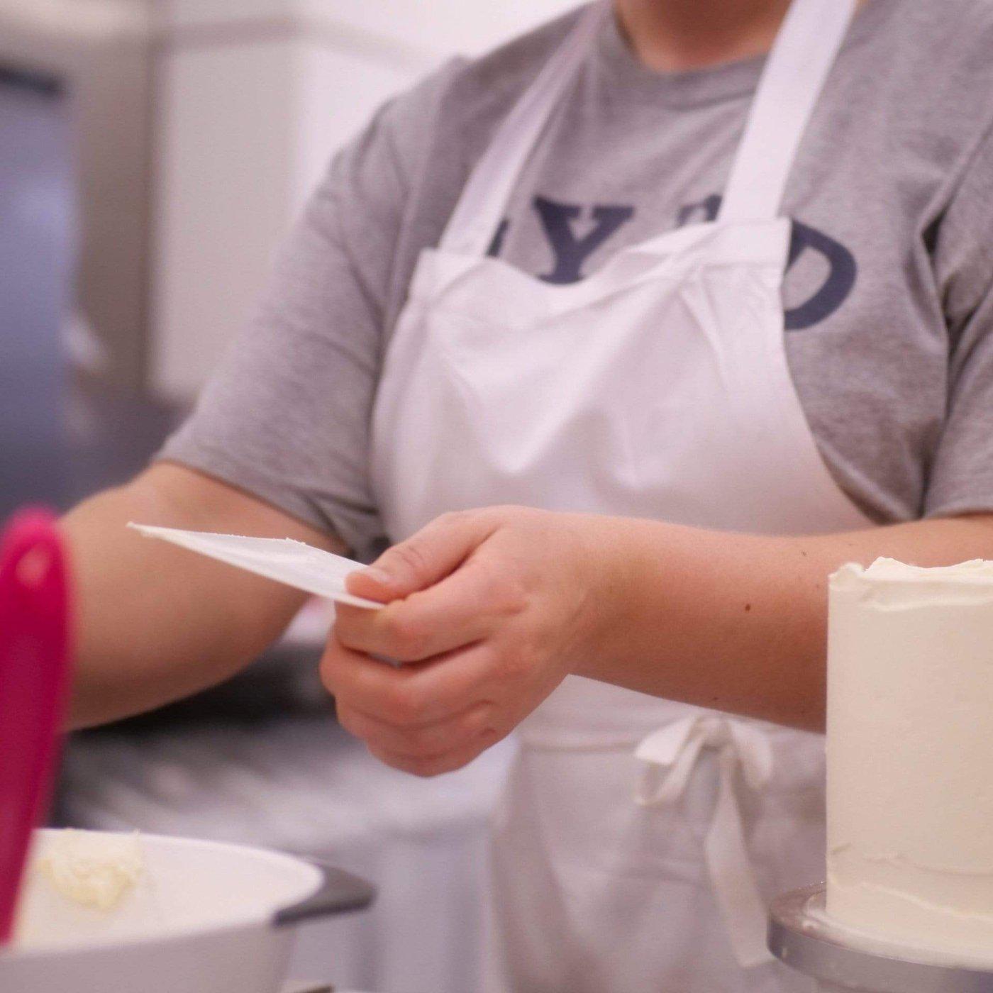 Person using a white Cake Scraper to smooth icing on a cake, wearing apron in a baking setting
