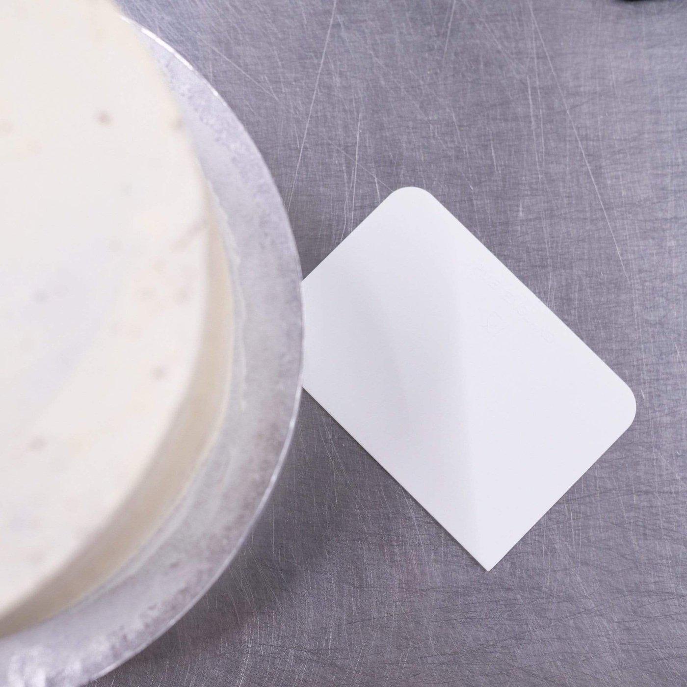 White cake scraper tool next to a frosted cake on a stainless steel surface for smooth edges